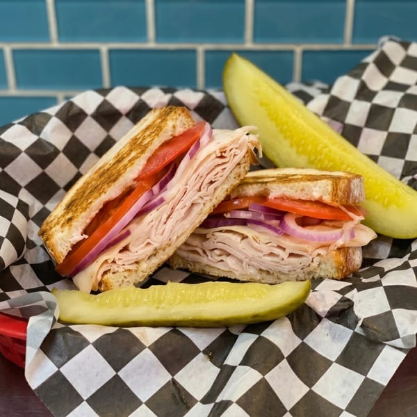 Toasted turkey sandwich with tomato and red onion, served with two pickle spears on checkered paper.
