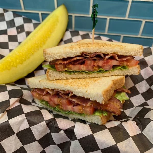 BLT sandwich with bacon, lettuce, tomato, and a pickle spear on checkered paper.