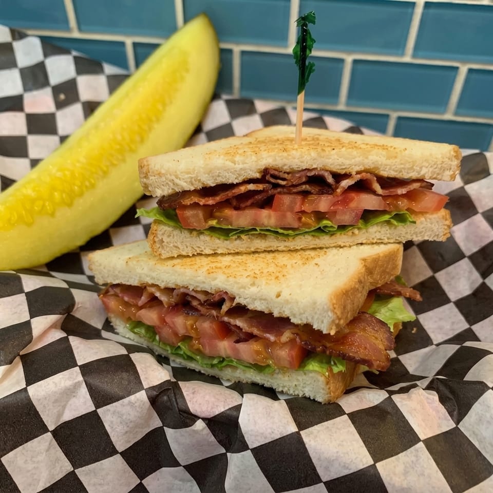 BLT sandwich with bacon, lettuce, tomato, and a pickle spear on checkered paper.