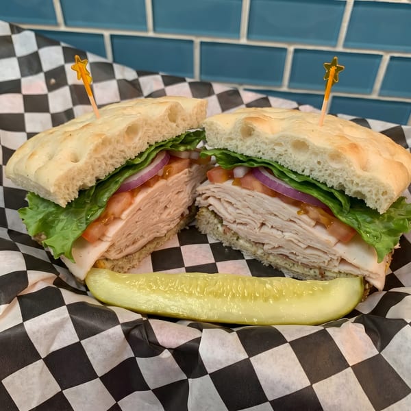 Turkey sandwich on focaccia bread with lettuce, tomato, and red onion, served with a pickle spear.