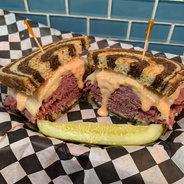 Reuben sandwich on marbled rye with melted cheese, sauce, and a pickle spear on checkered paper.