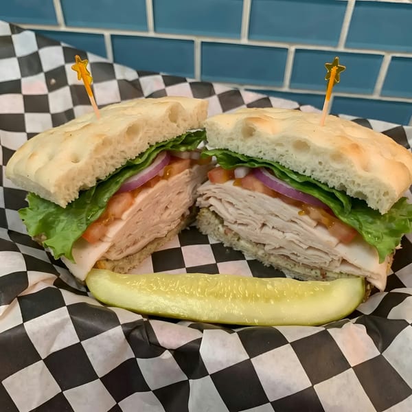 Turkey sandwich on focaccia with lettuce, tomato, and onion, served with a pickle spear.