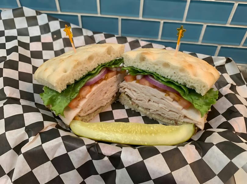 Turkey sandwich on focaccia with lettuce, tomato, and onion, served with a pickle spear.