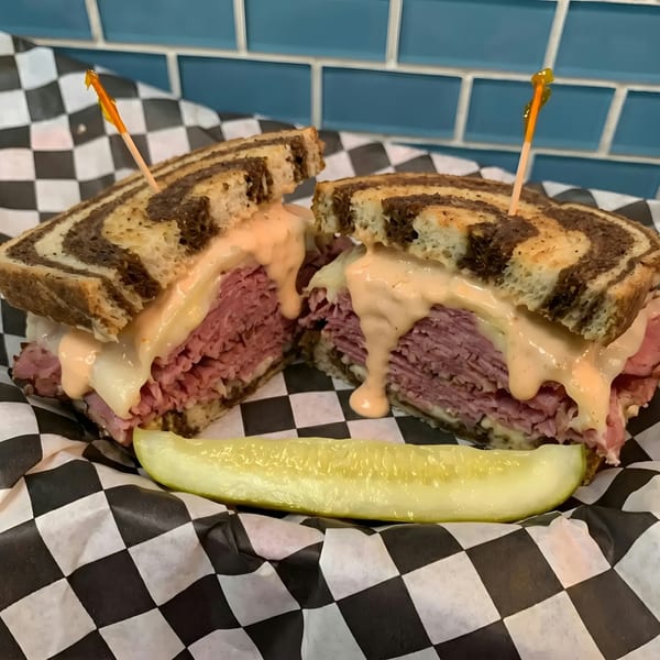 A reuben sandwich on marble rye with melted cheese and sauce, served with a pickle spear.