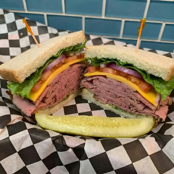 Roast beef sandwich with cheese, lettuce, tomato, and onion, served with a pickle spear.