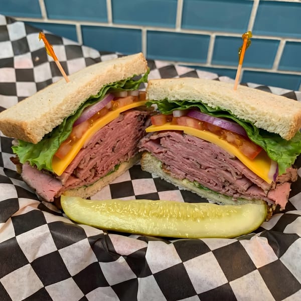 Roast beef sandwich with cheese, lettuce, tomato, and onion, served with a pickle spear.